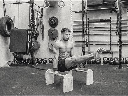 Mat Harvey on parallettes on the gym floor, with kettlebells, wall bars and the rig behind him