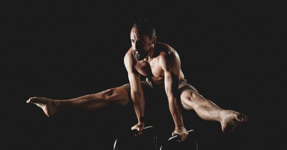 Mat Harvey holding a straddle on parallettes in a black studio