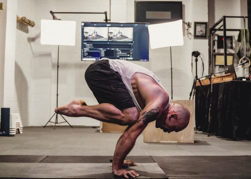 Mat Harvey in a crow pose on the gym floor, with the lights, camera and screens used to stream the classes behind him